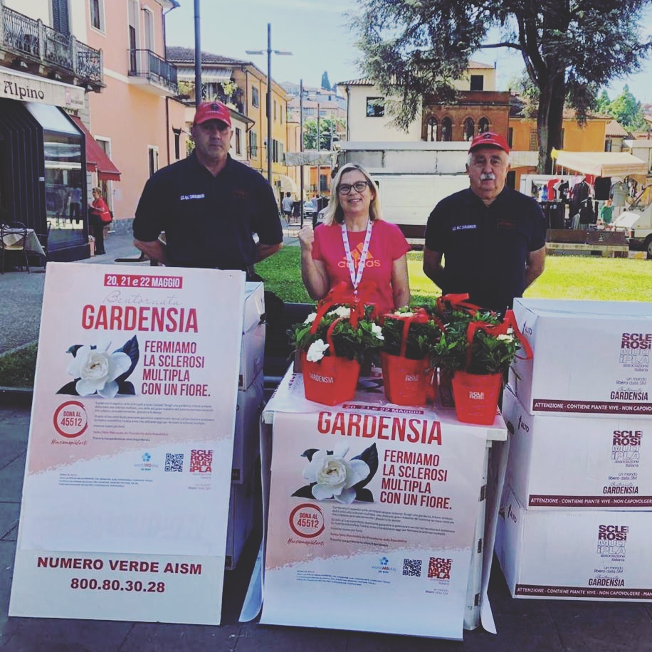 Il banchetto AISM in piazza a Barga, con 3 volontari che offrono una gardensia in cambio di un piccolo contributo a favore della ricerca contro la sclerosi multipla